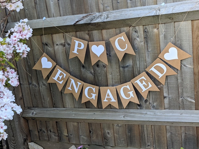 May include: A banner with the word "ENGAGED" and the initials "P C" with heart shapes. The banner is made of brown paper and white letters, hanging against a wooden fence. Pink flowers are visible on the left.