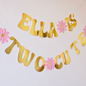 May include: Gold banner with the text "Ella is two cute" with pink paper flowers between each letter.