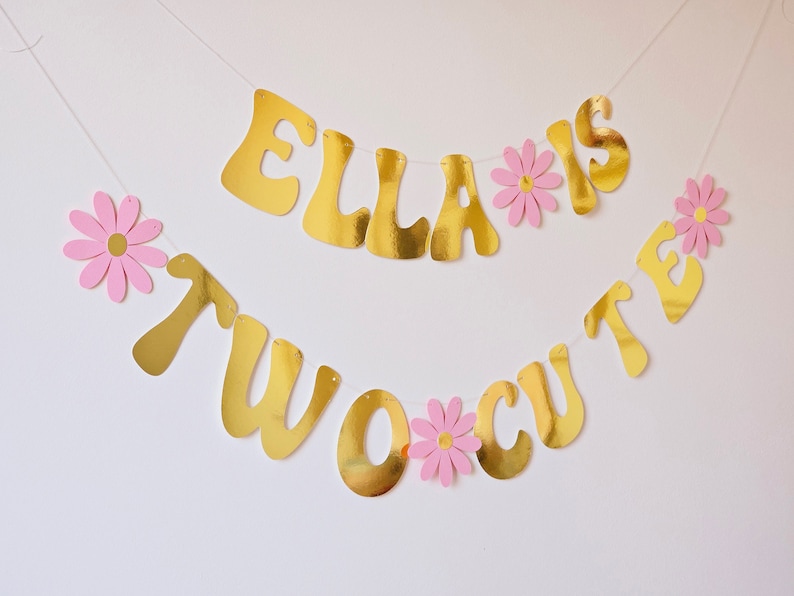 May include: Gold banner with the text "Ella is two cute" with pink paper flowers between each letter.