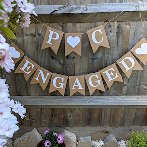 May include: A banner with the word "ENGAGED" and the initials "P C" in white letters on brown pennants. White heart shapes are also included. The banner is strung against a wooden fence, with flowers in the foreground.