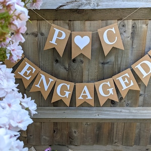 May include: A banner with the word "ENGAGED" and the initials "P C" displayed on brown pennants with white lettering and hearts. The banner is strung against a wooden fence, with pink and white flowers in the foreground.