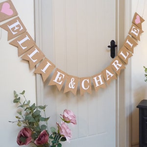 Pu&ograve; includere: Uno striscione di juta con i nomi "Ellie & Charlie" in lettere bianche e cuori rosa. Lo striscione &egrave; appeso a una porta bianca.