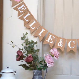 Pu&ograve; includere: Uno striscione di carta marrone con lettere bianche che compongono la scritta "ELLIE & C" con un cuore rosa sulla prima bandiera. Lo striscione &egrave; appeso a una parete bianca con un vaso di fiori rosa e bordeaux di fronte.
