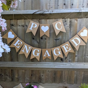 May include: A banner with the word "ENGAGED" and initials, made of brown paper pennants with white letters and hearts. The banner is strung on twine and hangs against a wooden fence, with pink flowers in the background.