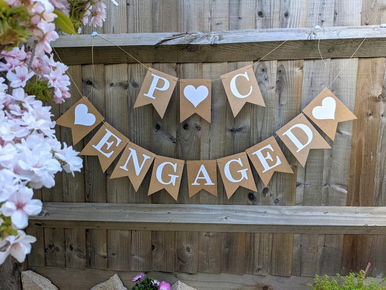 May include: A banner with the word "ENGAGED" and initials, made of brown paper pennants with white letters and hearts. The banner is strung on twine and hangs against a wooden fence, with pink flowers in the background.