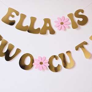 May include: Gold banner with the text "Ella is two cute" and pink paper flowers. The banner is strung together with white string.