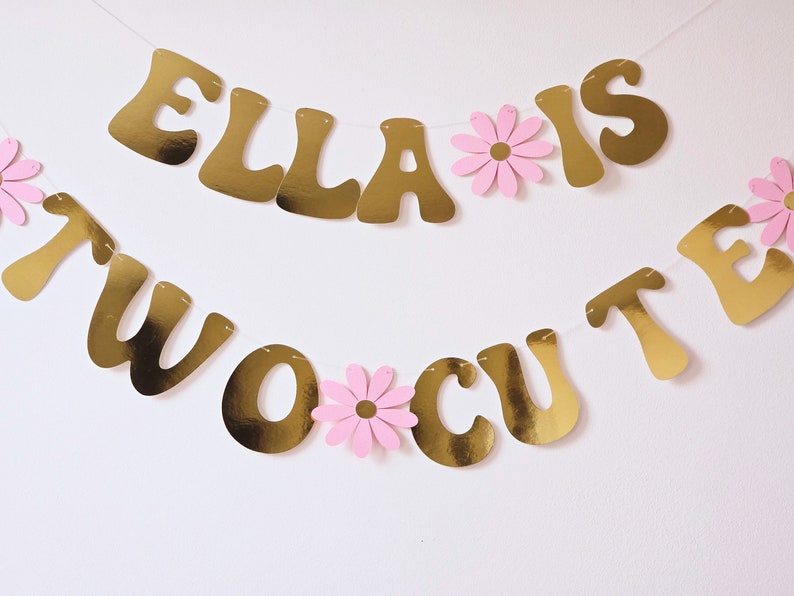 May include: Gold banner with the text "Ella is two cute" and pink paper flowers. The banner is strung together with white string.