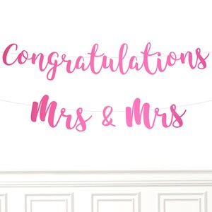 May include: A pink banner with the words "Congratulations Mrs & Mrs" in a cursive font. The banner is strung across a white background, suitable for a wedding or engagement celebration.