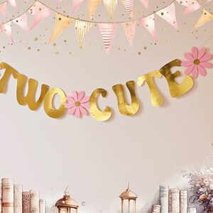 May include: Gold foil banner with the words "TWO CUTE" in a cursive font. The banner is decorated with pink paper flowers.