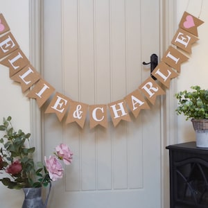 Pu&ograve; includere: Uno striscione di juta con i nomi "Ellie & Charlie" in lettere bianche e cuori rosa ad ogni estremit&agrave;. Lo striscione &egrave; appeso a una porta bianca.
