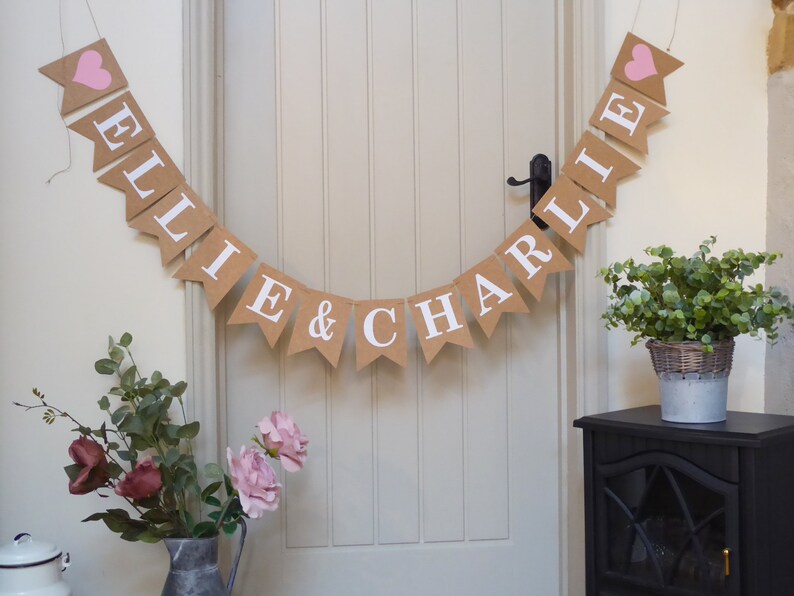Pu&ograve; includere: Uno striscione di juta con i nomi "Ellie & Charlie" in lettere bianche e cuori rosa ad ogni estremit&agrave;. Lo striscione &egrave; appeso a una porta bianca.