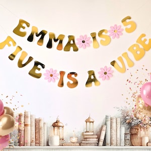May include: Gold banner with the text "Emma is 5" and "Five is a vibe" with pink flowers between each letter. The banner is hanging on a white wall. There are pink and gold balloons on a shelf with books and lanterns.