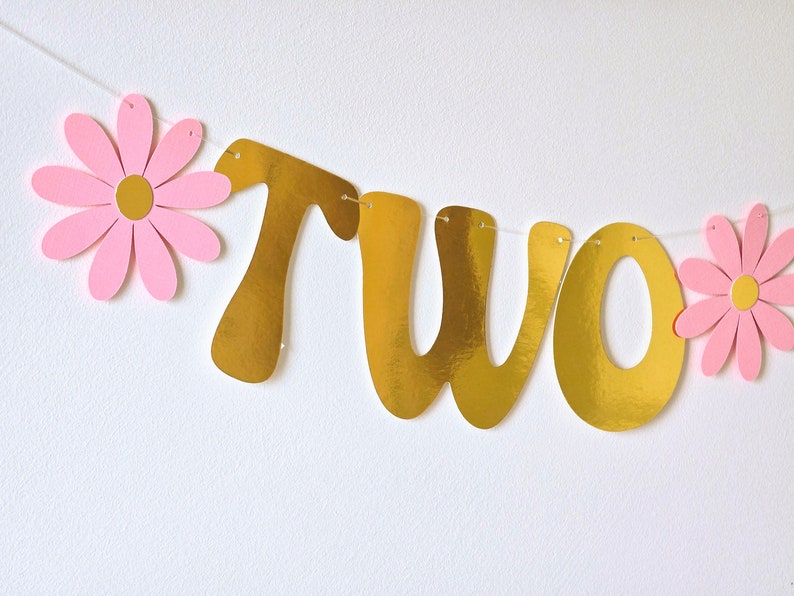 May include: Gold glitter "two" banner with pink paper flowers. The banner is strung with white thread.