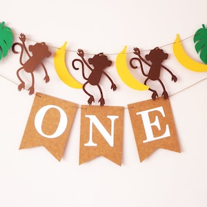 Peut inclure: Une bannière en papier brun avec le mot "ONE" en lettres blanches. La bannière est décorée de feuilles de palmier vertes, de singes bruns et de bananes jaunes.