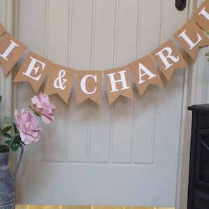 Pu&ograve; includere: Uno striscione di carta marrone con lettere bianche che compongono la scritta "Ellie & Charlie" appeso a una porta bianca. Lo striscione &egrave; decorato con rose rosa.