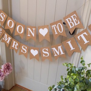 Pu&ograve; includere: Uno striscione di carta marrone con lettere bianche che recita "Mr & Mrs Soon to be Smith" con cuori bianchi tra le parole "Soon" e "Smith".