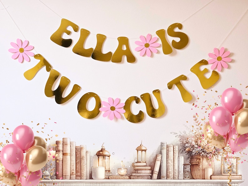 May include: Gold banner with the text "Ella is two cute" and pink paper flowers. The banner is hanging on a white wall. There are pink and gold balloons and confetti on a shelf below the banner.