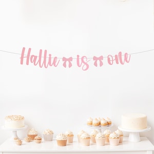 May include: Pink banner with the text "Hallie is one" with pink bows on either side of the word "is". The banner is hanging above a table with cupcakes and cakes.