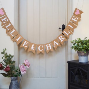 Pu&ograve; includere: Uno striscione di juta con cuori rosa e i nomi "Ellie & Charlie" in lettere bianche. Lo striscione &egrave; appeso a una porta bianca con una maniglia nera. A destra della porta ci sono piante verdi in un vaso su un supporto nero.