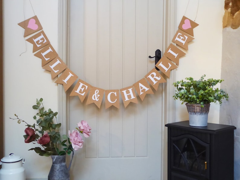 Pu&ograve; includere: Uno striscione di juta con cuori rosa e i nomi "Ellie & Charlie" in lettere bianche. Lo striscione &egrave; appeso a una porta bianca con una maniglia nera. A destra della porta ci sono piante verdi in un vaso su un supporto nero.