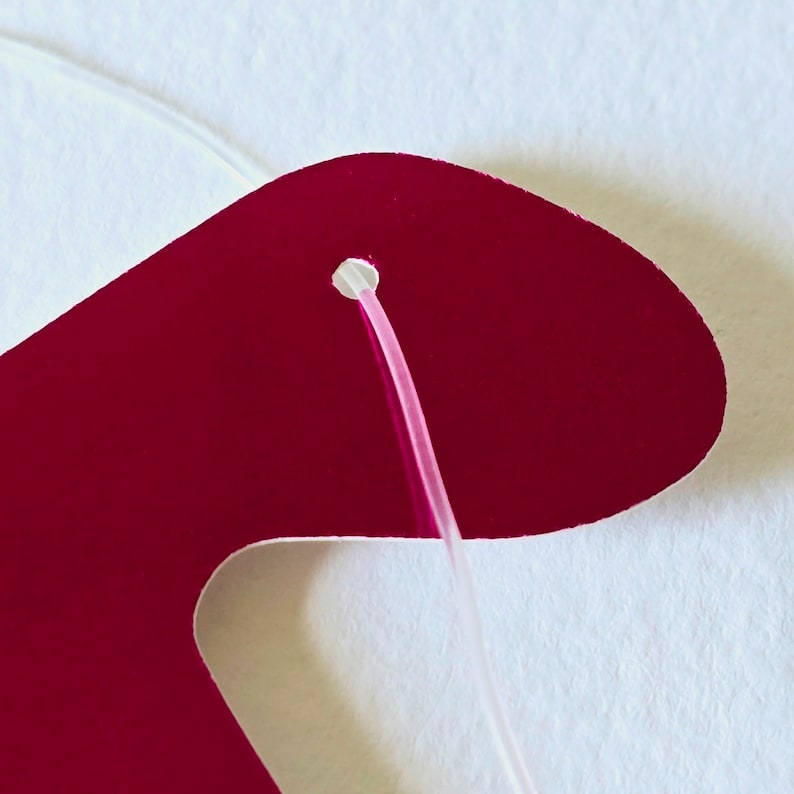 May include: A close-up of a red paper cutout with a hole in the top. A white string is threaded through the hole.