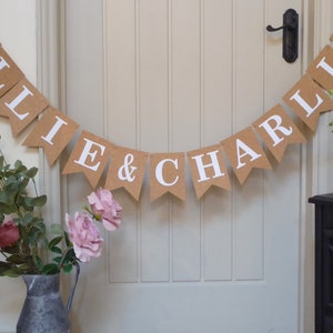 Pu&ograve; includere: Uno striscione di juta marrone con lettere bianche che compongono "Ellie & Charlie" con cuori rosa ad ogni estremit&agrave;. Lo striscione &egrave; appeso a una porta bianca.