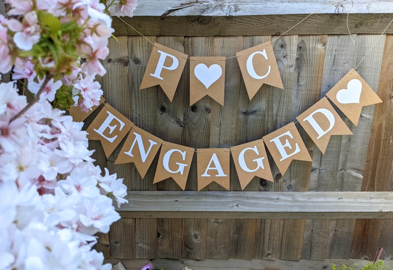 May include: A banner with the word "ENGAGED" and the initials "P C" displayed on brown pennants with white lettering and hearts. The banner is strung against a wooden fence, with pink and white flowers in the foreground.