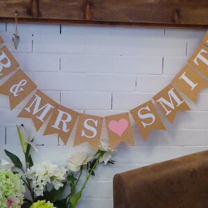 Pu&ograve; includere: Uno striscione di carta marrone con lettere bianche che recita "MR & MRS SMITH" con un cuore rosa tra le ultime due parole. Lo striscione &egrave; appeso a un muro di mattoni bianchi.