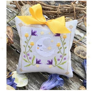 May include: A cross-stitch design named "Light In The Darkness" showing a light blue pillow with a crescent moon and star pattern. It has purple flowers, yellow accents, and a yellow bow. The design is by Maja (Mátyás) Merész.
