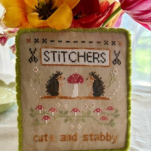 May include: A cross-stitch embroidery piece with a green border. The design features two hedgehogs under a red mushroom with the text "STITCHERS" above them and "cute and stabby" below.