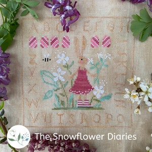 Może przedstawiać: Wzór do haftu krzyżykowego przedstawiający różowego króliczka trzymającego kwiat w ogrodzie z białymi kwiatami i zielonymi liśćmi. Wzór zawiera tekst "The Snowflower Diaries".