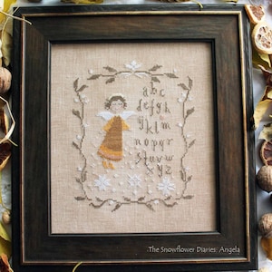Peut inclure: Une broderie encadrée représentant un ange en robe moutarde, entouré d'un alphabet et d'une bordure florale. L'œuvre est posée sur un fond beige et encadrée dans un cadre en bois brun foncé. Le texte "The Snowflower Diaries: Angela" est visible.
