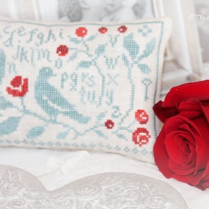 Puede incluir: Una almohada blanca con un pájaro azul y flores rojas bordadas. La almohada tiene el alfabeto bordado en una fuente caprichosa. La almohada está sentada sobre una superficie blanca con una rosa roja.