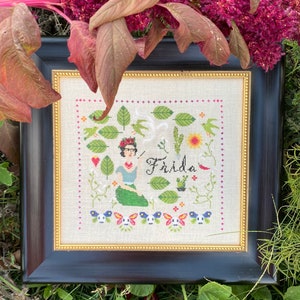 Pode incluir: Bordado de ponto cruz emoldurado com Frida Kahlo com um pincel, cercada por flores, folhas e a palavra "Frida".