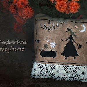 Könnte beinhalten: Ein dekoratives Kissen mit Kreuzstichmuster, das eine Hexe, einen Kessel und einen Kronleuchter zeigt. Das Kissen hat eine cremefarbene Spitzentrimmung und den Text "The Snowflower Diaries: Persephone". Der Hintergrund zeigt orangefarbene Blumen.