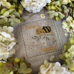 May include: Cross-stitch embroidery with a bee and flowers, surrounded by white and green hydrangeas. The embroidery reads "Bee Happy!"