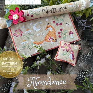 Op de afbeelding: Een kruissteekpatroon met een vos, bloemen en het woord "Natuur". Het ontwerp bevat een klein kussen en een opgerold stuk met het woord "Natuur". De tekst "Abundance" is ook zichtbaar.