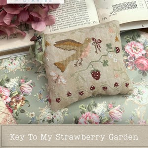 Może przedstawiać: Poduszka do haftu krzyżykowego z ptakiem, truskawkami i kwiatowymi detalami. Projekt zawiera tekst "Key To My Strawberry Garden" i "a cross stitch design by Maja Merész."