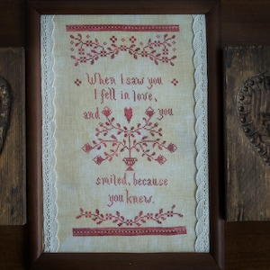 Può includere: Un ricamo a punto croce incorniciato con sfondo bianco e ricamo rosso. Il ricamo presenta un motivo floreale a forma di cuore e il testo "When I saw you I fell in love, and you smiled, because you knew."