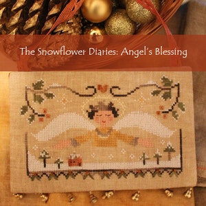 May include: A cross-stitch sampler featuring a white angel with wings outstretched, flying over a snowy landscape with trees and a small house. The sampler is framed with a brown border and has a fringe of gold beads at the bottom. The title "The Snowflower Diaries: Angel's Blessing" is displayed above the sampler.