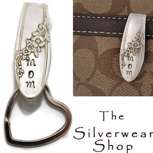 May include: Silver spoon keychain with a heart-shaped ring and the word "MOM" engraved on it. The spoon has a floral design and is attached to a brown leather purse.