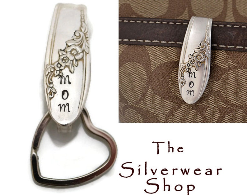 May include: Silver spoon keychain with a heart-shaped ring and the word "MOM" engraved on it. The spoon has a floral design and is attached to a brown leather purse.