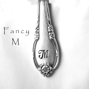 May include: Silver spoon handle with a floral design and the letter "M" engraved on it. The spoon handle is on a white background with the words "Fancy M" written in black.