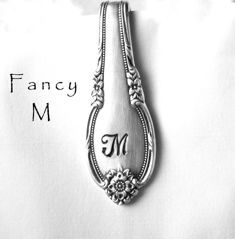 May include: Silver spoon handle with a floral design and the letter "M" engraved on it. The spoon handle is on a white background with the words "Fancy M" written in black.