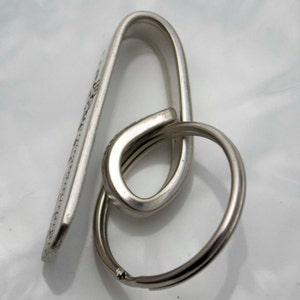 May include: Silver key chain with a hook and a ring. The hook is shaped like a loop and has a flat surface with a design.