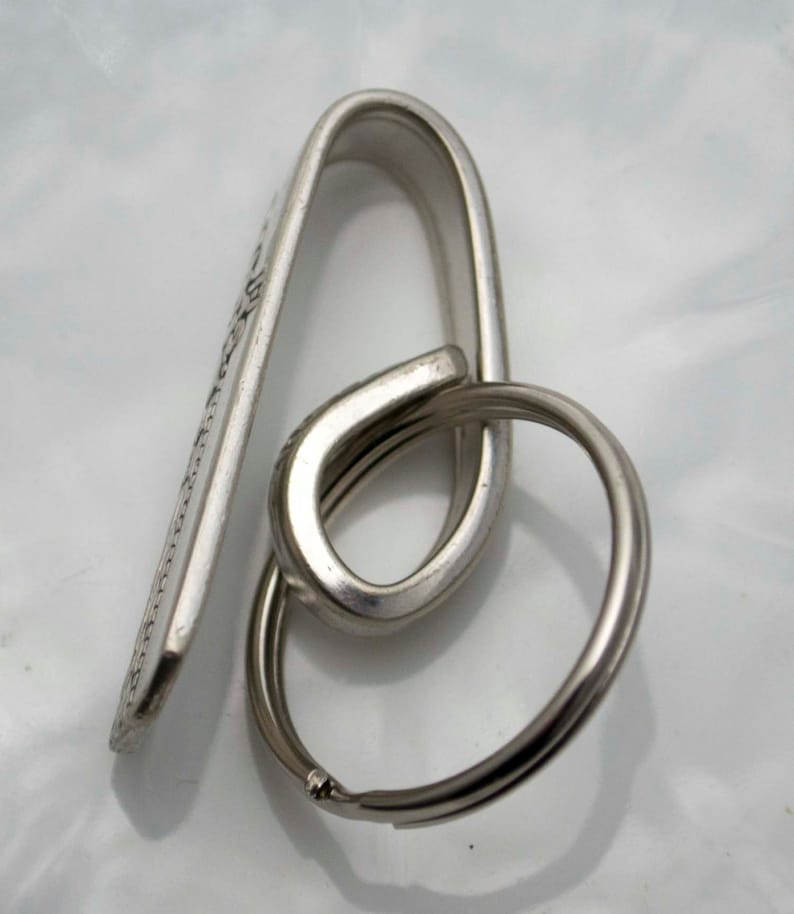 May include: Silver key chain with a hook and a ring. The hook is shaped like a loop and has a flat surface with a design.