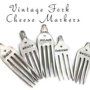 May include: Set of six vintage silver forks with different cheese names engraved on them: Swiss, Gouda, Asiago, Cheddar, Smelly.  The forks are arranged in a semi-circle on a white background.