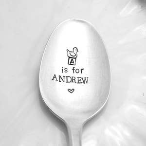 May include: A silver spoon with the text "A is for ANDREW" engraved on it. A small heart is engraved below the text. A cartoon duck is engraved above the text, sitting on top of a block with the letter "A" on it.
