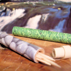 May include: Two round, cylindrical toothpicks holders. One is white with a pattern of gray stones, the other is green with a pattern of green grass. Both holders have a white cap on one end and are filled with wooden toothpicks.