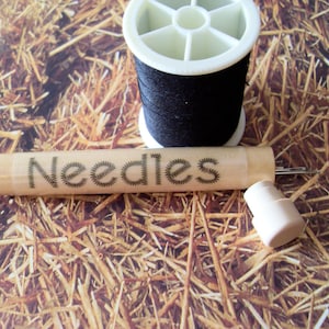 May include: A wooden case with the word "Needles" printed on it. The case contains a set of needles and a spool of black thread.